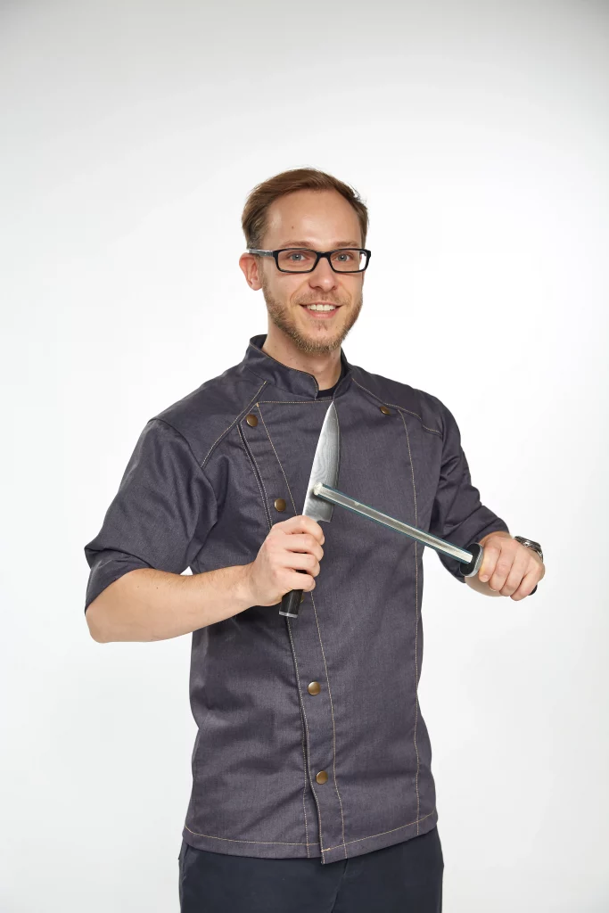 Einen Koch, der ein Messer an einem Wetzstahl schärft. Der Koch trägt eine dunkelblaue Kochjacke.