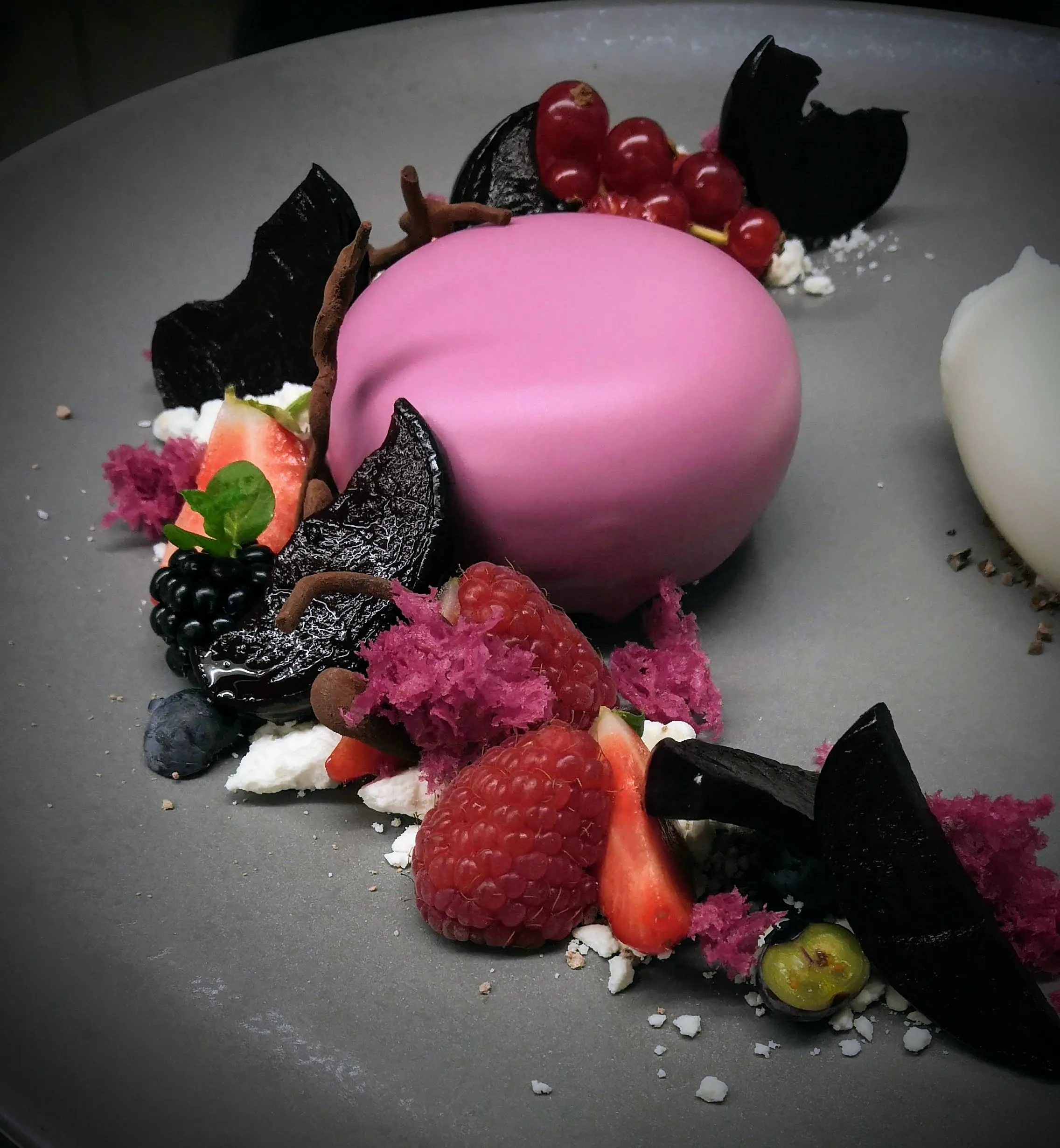 Ein exquisites Dessert auf einem grauen Teller. In der Mitte des Tellers befindet sich eine rosa Mousse, umgeben von frischen Beeren wie Erdbeeren, Himbeeren, Brombeeren und Johannisbeeren. Um die Beeren herum sind Schokoladensplitter kunstvoll platziert, die einen dunklen Kontrast zu den lebendigen Farben der Früchte und der rosa Mousse bilden. Zerbröselte rosa Kuchen- oder Keksbrösel sind um das Dessert herum gestreut und verbessern die visuelle Wirkung. Eine Portion weiße Sahne ist auf der rechten Seite des Tellers sichtbar und sorgt für einen weichen Texturkontrast.
