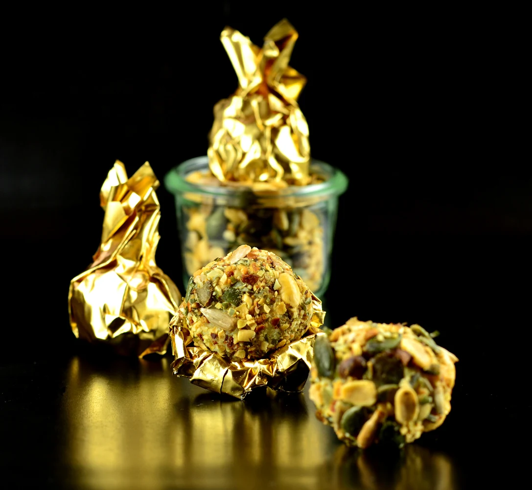 Drei goldverpackte Pralinen und zwei unverpackte Pralinen mit Nussstücken, die auf einer reflektierenden dunklen Oberfläche platziert sind. Die goldverpackten Pralinen haben eine glänzende, geknitterte Textur und sind an den Enden verdreht. Die unverpackten Pralinen zeigen eine Mischung aus Nüssen, die in ihnen eingebettet sind. Ein kleiner Glasbehälter, der mit mehr goldverpackten Pralinen gefüllt ist, ist im Hintergrund sichtbar. Die dunkle Oberfläche und der Hintergrund bilden einen starken Kontrast zur goldenen Farbe der Bonbonverpackungen, wodurch sie deutlich hervorstechen.