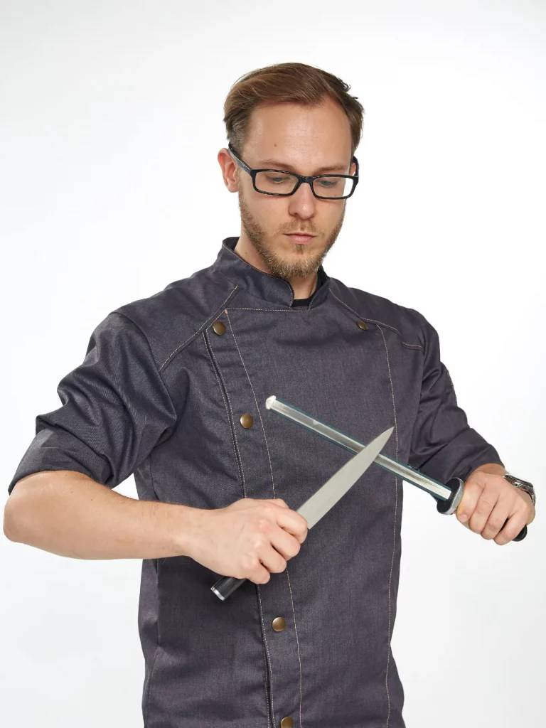 Einen Koch, der ein Messer an einem Wetzstahl schärft. Der Koch trägt eine dunkelblaue Kochjacke 1. Das Schärfen von Messern ist ein wichtiger Bestandteil der Arbeit eines Kochs, da scharfe Messer die Arbeit erleichtern und sicherer machen 2. Ein Wetzstahl ist ein Werkzeug, das verwendet wird, um die Schärfe von Messern wiederherzustellen.