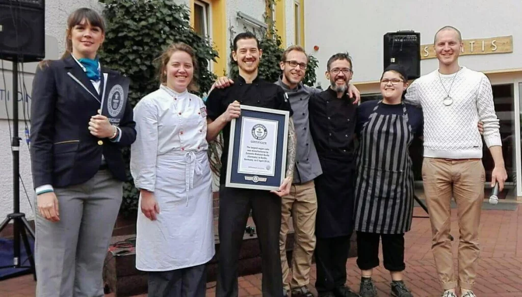 Eine Gruppe von sieben Personen, die nebeneinander im Freien stehen. Sie tragen verschiedene Kleidungsstücke, darunter Kochuniformen und Freizeitkleidung. Eine Person hält prominent vor der Gruppe ein Zertifikat des Guinness-Buchs der Rekorde für den größten Veganen Kuchen der Welt . Die Einstellung scheint eine Outdoor-Veranstaltung zu sein, mit einem Lautsprechersystem im Hintergrund und einem Teil eines Gebäudes und einer Beschilderung ebenfalls sichtbar. Die Stimmung scheint feierlich zu sein, da sie eine Anerkennung erhalten haben, die durch das Zertifikat angezeigt wird.