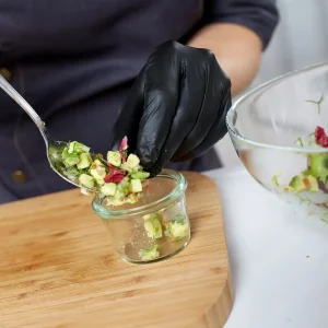 Ein professioneller Koch bereitet einen gesunden Salat zu. Der Koch trägt schwarze Handschuhe und verwendet einen silbernen Löffel, um den Salat aus einer klaren Schüssel in ein kleines Glasgefäß zu geben. Der Salat besteht aus gehacktem Gemüse in verschiedenen Farben, was auf eine Vielzahl von Zutaten hinweist. Der Koch arbeitet auf einem hölzernen Schneidebrett, was auf eine Kücheneinstellung hinweist